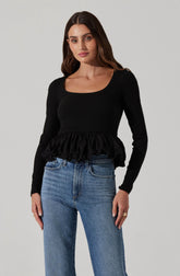 A woman is wearing a black, long-sleeved top with a square neckline. The top features a ruffled hem that adds a decorative and feminine touch. She pairs it with high-waisted blue jeans.